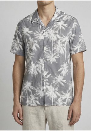 Homme portant une chemise grise à manches courtes boutonnée avec un imprimé de palmiers blancs et un pantalon de couleur claire, debout devant un fond gris.