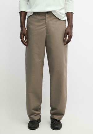 Mand iført løsne beige bukser, sorte sko og en hvid langærmet skjorte, stående mod en ensfarvet baggrund.