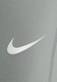 Leggings de sport Nike en tissu gris clair, avec un logo en Swoosh argent métallique, une surface texturée et des coutures visibles.