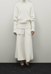 Pull en tricot blanc avec un col montant, superposé à une chemise blanche, associé à une jupe blanche fluide et des chaussures noires à talons pointus, sur un fond neutre.