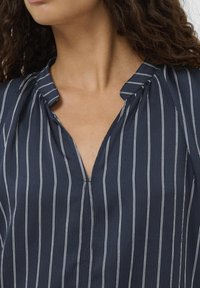 Tengerészkék, rövid ujjú blúz fehér, függőleges csíkokkal, texturált anyagból, galléros V-nyakkal és finom ráncolással a vállaknál.