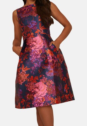 Woman wearing a sleeveless, knee-length dress with a vibrant red, pink, and purple floral pattern on a dark blue background, hands in pockets.