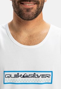 Biela bavlnená tielko s modro orámovanou grafikou, na ktorej je text "Quiksilver" v čiernej farbe a ikona hory, umiestnená na oblasti hrudníka.