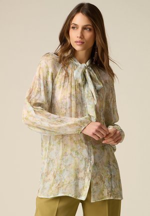 Kobieta ubrana w przezroczystą, długorękawową bluzkę w kwiaty z kokardą wiązaną na szyi, w zestawieniu z oliwkowozielonymi spodniami, na jednolitym tle.