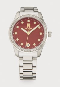 Nerezová hodinka s kulatým pouzdrem, červeným ciferníkem, zlatými akcenty, krystalovými indexy a stříbrným odkazem. Detaily na lunetě ve stylu diamantu.