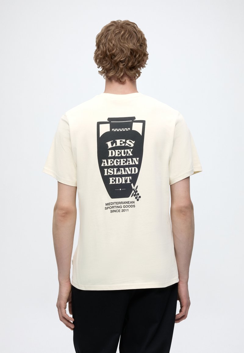 Άτομο με σγουρά μαλλιά φοράει ένα κρεμ T-shirt με μαύρο σχέδιο ενός βάζου και κείμενο για το Αιγαιοπελαγίτικο νησί στην πλάτη, στέκεται μπροστά σε απλό φόντο.