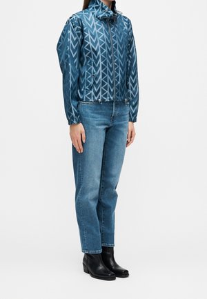 Kvinde iført en blå mønstret jakke med lynlås, lige blå jeans og sorte ankelstøvler, stående med hænder afslappet ned langs siderne.