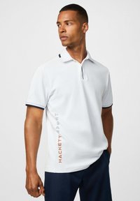 Muž v bielom polokošeli s čiernym lemovaním a textom "Hackett Sport" na boku, v tmavomodrých nohaviciach, pozerajúci sa doľava.