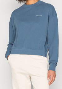 Sudadera de algodón azul con cuello redondo, puños y dobladillo acanalados. Presenta un pequeño logo en el pecho. Combinada con pantalones de color claro.
