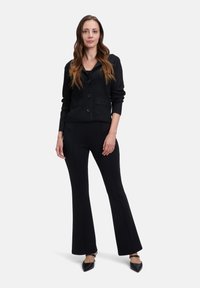 Svart strukturerad cardigan med V-ringning, knappstängning och två fickor, kombinerad med svarta utsvängda byxor och svarta skor med spetsiga tår.