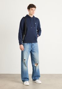 Felpa con cappuccio blu navy con lacci e tasca frontale, abbinata a jeans in denim strappati blu chiaro e scarpe da ginnastica bianche. Borsa nera appesa alla spalla.