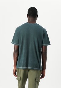 T-shirt in cotone verde scuro con vestibilità rilassata, maniche corte e scollatura a girocollo. Presenta cuciture visibili lungo l'orlo e le maniche.
