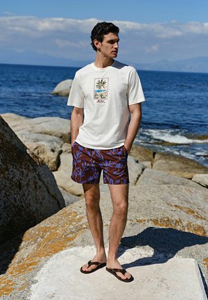 Bărbat purtând tricou alb cu imprimeu de palmier, pantaloni scurți florali maro și papuci, stând pe stânci lângă ocean într-o zi însorită.