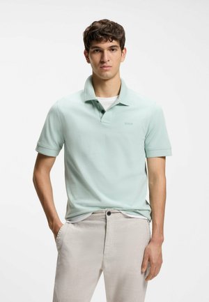 Jeune homme portant un polo vert clair et un pantalon beige clair, debout avec une main dans la poche, sur fond blanc uni.