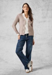 Helles rosa geripptes Cardigan mit Knöpfen, über einem weißen Hemd getragen. Kombiniert mit lockeren blauen Denim-Jeans und weißen Sneakers mit schwarzen Akzenten.