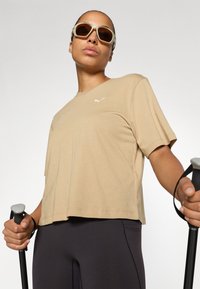 Camiseta de manga corta beige con un pequeño logo blanco en el pecho, combinada con leggings deportivos oscuros. La persona sostiene bastones de senderismo.