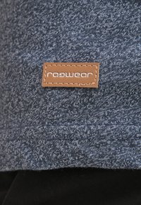 Brun rektangulær stofetiket med hvid "ragwear" tekst syet på blågråt strikket stof nær kanten.