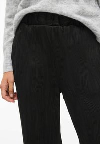 Pantalon noir plissé avec une finition texturée et une taille élastique, associé à un pull gris clair. Main partiellement visible.