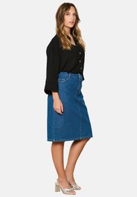 Denim A-linija krilo v srednje modri barvi, dolžine do kolen, v kombinaciji s črno srajco na gumbe. Srebrni sandali s peto in teksturiranimi trakovi.