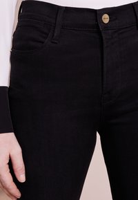 Pantalones vaqueros de mezclilla negra con un diseño ajustado, con cierre de botón dorado y bolsillos delanteros, textura suave y detalles de costura mínimos.