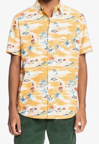 Kortärmad skjorta i gul tyg med ett tropiskt mönster som har palmträd, segelbåtar och blommiga detaljer. Enfärgad bröstficka.