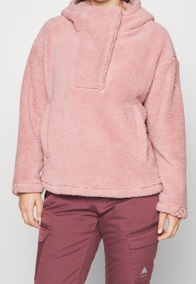 Personne portant un sweat à capuche en polaire rose et un pantalon cargo bordeaux avec poches zippées et à rabat, se tenant devant un fond uni.