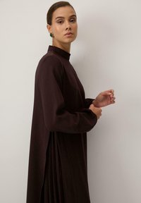 Rochie maro, cu mâneci lungi și guler înalt, cu un design plisat, realizată dintr-un material moale. Modelul se află în profil, într-un fundal neutru.