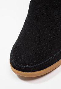 Czarne zamszowe buty o perforowanej cholewce, zaokrąglonym nosku i jasno-brązowej gumowej podeszwie. Powierzchnia ma miękką teksturę i elegancki design.