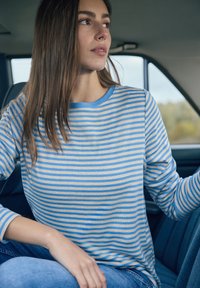 Chemise à manches longues rayée en bleu clair et crème, fabriquée en tissu doux. Elle présente un col rond et une coupe décontractée, portée avec un jean en denim.