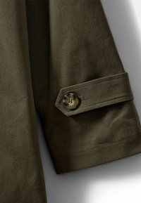 Tessuto di camicia verde oliva scuro con una trama liscia, caratterizzato da un polsino abbottonato con un bottone circolare e decorato.