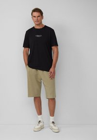 Zwarte katoenen t-shirt met een klein logo op de borst, gecombineerd met beige shorts. Model draagt witte sneakers en enkelsokken. Eenvoudige achtergrond.