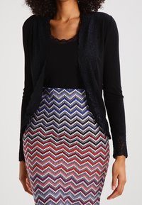 Černý kardigan s dlouhým rukávem a krajkovým lemováním, doplněný sukní s vícenásobným vzorem cik-cak v červených, modrých a stříbrných tónech.