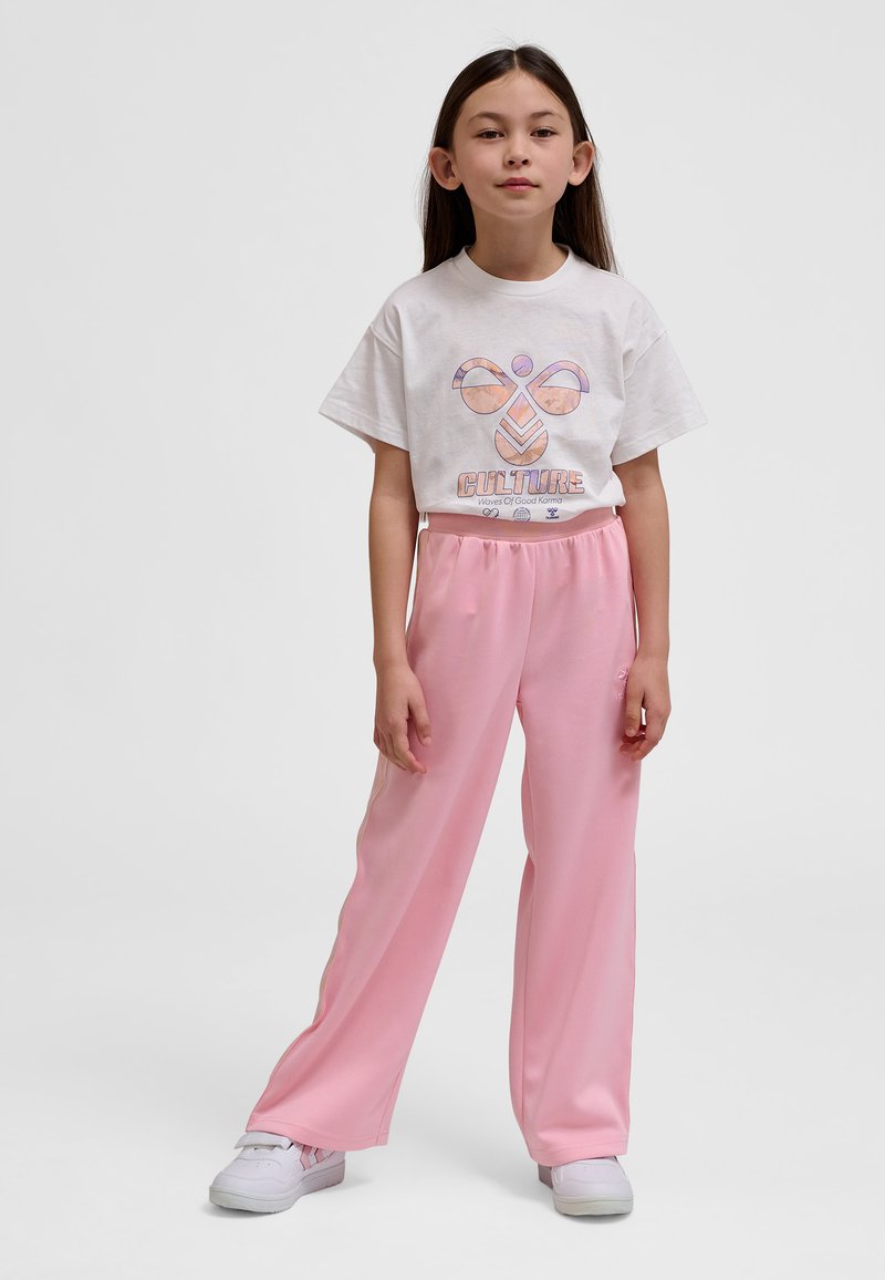 Młoda dziewczyna stoi, ubrana w białą krótka koszulkę z nadrukiem oraz różowe luźne spodnie, białe trampki, neutralne szare tło.