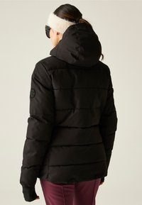 Vrouw in een zwarte gewatteerde winterjas met capuchon, een crèmekleurige gebreide hoofdband, zonnebril en paarse broek, met haar rug naar de camera gekeerd tegen een effen achtergrond.