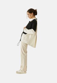 Chaqueta ligera con la parte superior negra y la inferior crema, que cuenta con un bolsillo bordado y cierre con cordón, combinada con pantalones beiges y zapatillas.