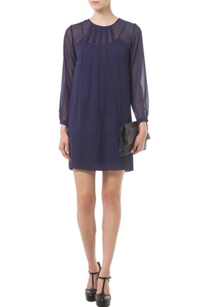 Mörkblå klänning med långa genomskinliga ärmar, prickigt mönster, rund halsringning och veck vid bysten. Modellen håller en svart clutch.