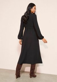 Dlhé čierne šaty s priliehavým pásom, dlhými rukávmi a rozšírenou sukňou. Nosí sa s hnedými vysokými čižmami na vysokom opätku po kolená. Mäkká, štruktúrovaná tkanina.