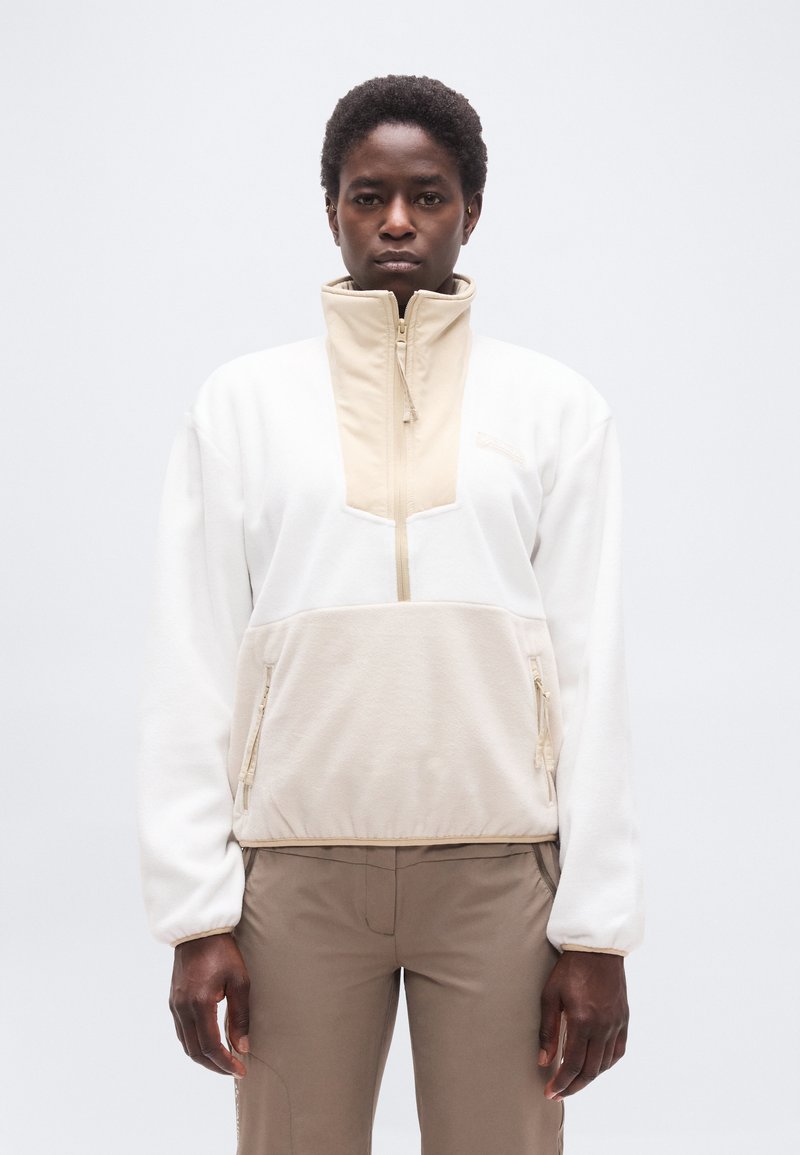 Άτομο που κοιτάζει μπροστά, φορώντας μπεζ και λευκό fleece σακάκι με φερμουάρ και ανοιχτό καφέ παντελόνι, με φόντο απλό λευκό.