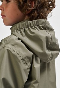 Kurtka z kapturem w kolorze khaki, wykonana z lekkiego, gładkiego materiału. Posiada marszczony dekolt i luźny fason z minimalną ilością przeszyć.