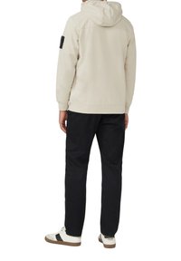 Felpa beige chiaro con cappuccio, caratterizzata da una patch nera sul braccio sinistro. Abbinata a pantaloni neri e sneakers bianche con dettagli marroni.