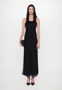Rochie maxi neagră, fără mâneci, realizată dintr-un material fin și ușor. Include o fante laterală, asortată cu o poșetă neagră și pantofi cu vârf ascuțit.