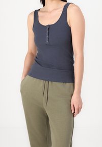 Kvinna klädd i en mörkgrå ribbad linne med knappar och olivgröna dragsko-byxor, stående med en hand i fickan.