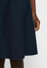 Robe tricotée marine avec un ourlet évasé, tissu texturé, et gros plan d'une main sur le bord de la jupe, mettant en valeur le matériau et le design.