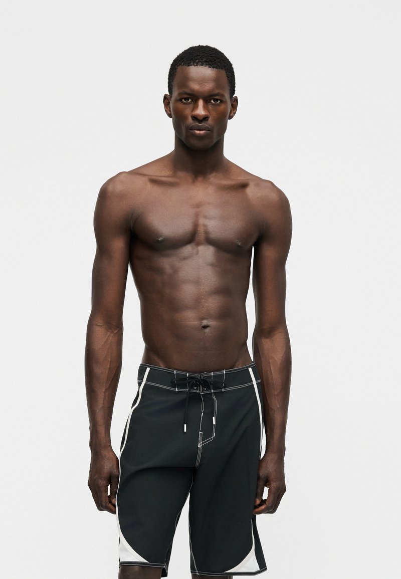 Αθλητικός άνδρας με σκουρόχρωμο δέρμα που φοράει μαύρα και λευκά μαγιό, στέκεται μπροστά σε απλό ανοιχτό γκρι φόντο με ουδέτερη έκφραση.