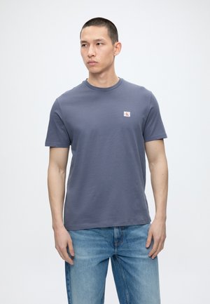 Άνδρας που στέκεται, φορά ένα μπλε κοντομάνικο μπλουζάκι με μικρό λογότυπο στο στήθος και ανοιχτό μπλε τζιν, σε λευκό φόντο.