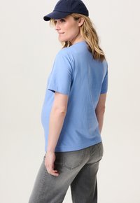 Lichtblauw T-shirt met korte mouwen van katoen, gedragen met donkergrijze jeans. Een marineblauwe pet rust op het hoofd van het model, met blootgestelde schouders.