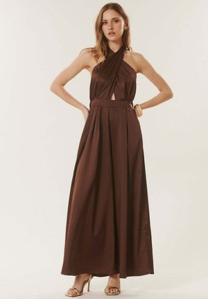 Femme portant une robe maxi sans manches à col licou brun foncé avec des plis et des sandales beiges à talons hauts, debout les mains sur les hanches.