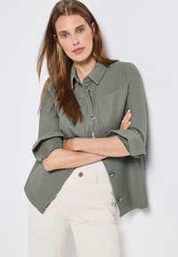 Jasnozielona koszula na guziki z kołnierzykiem, podwiniętymi rękawami i kieszonką z przodu. Miękka tekstura, lekko oversize'owy krój, zestawiona z kremowymi spodniami.