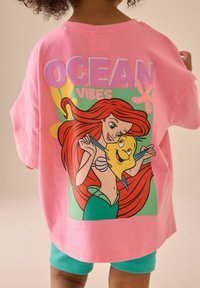 Lyserød bomulds t-shirt med et grafisk afbildning af en tegneseriefigur med bølgende rødt hår og en gul fisk, teksten lyder "OCEAN VIBES".