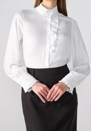 Mujer con blusa blanca de manga larga con volantes en el frente y los puños, combinada con una falda negra de talle alto, con las manos entrelazadas al frente.